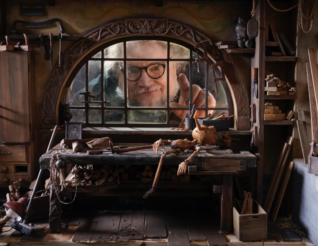 "Gullermo del Toro's Pinocchio" promo image featuring the director himself looking through a window at a small Pinocchio puppet.